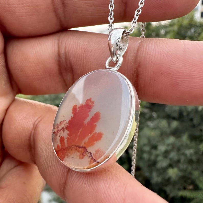 May include: Oval pendant necklace featuring a translucent stone with a red and brown dendritic pattern. The stone is set in a silver-colored metal bezel and hangs from a delicate silver chain. The pendant is held by a hand.
