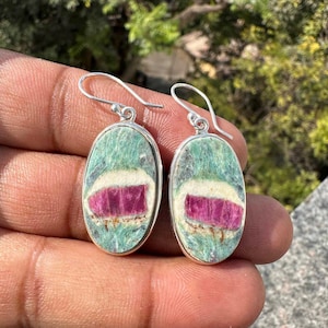 Peut inclure: Boucles d'oreilles ovales avec crochets en argent. Chaque boucle présente une pierre verte et blanche avec un centre magenta. Les boucles d'oreilles sont présentées sur un fond naturel flou.