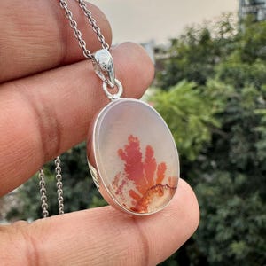 May include: Oval pendant with a silver chain. The pendant features a translucent stone with a red and orange dendritic pattern. The stone is set in a silver bezel. The chain is a delicate silver link chain.