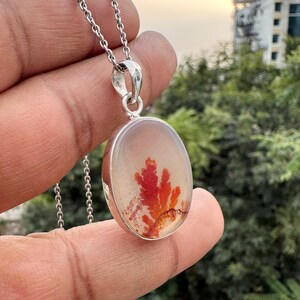 May include: Silver pendant necklace featuring an oval-shaped, translucent stone with a vibrant red and orange dendritic pattern. The pendant is suspended from a delicate silver chain, showcasing a natural, artistic design. The necklace is a unique piece of jewelry.