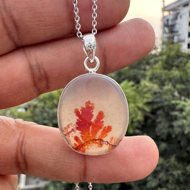 May include: Silver pendant necklace featuring a round, clear stone with a vibrant red and orange dendritic inclusion. The pendant hangs from a delicate silver chain. The inclusion resembles a fiery, branching pattern.