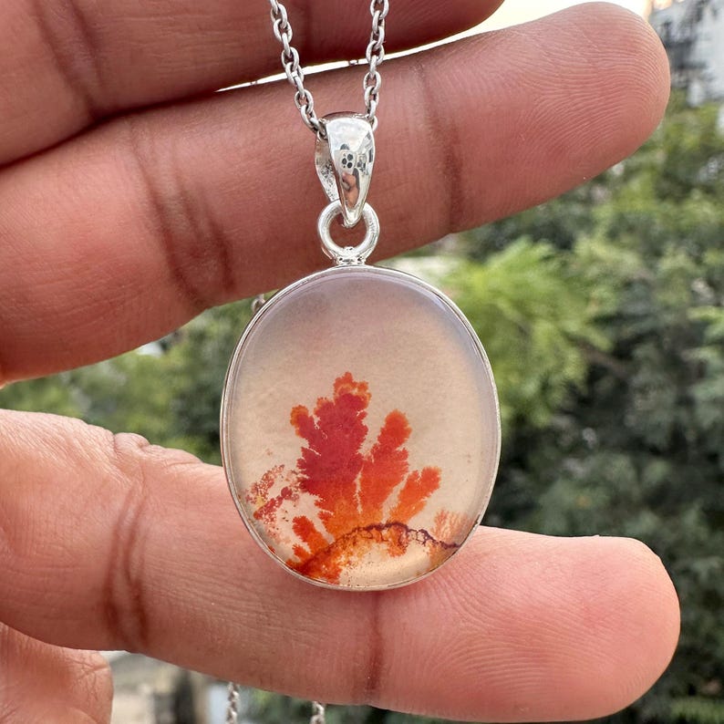 May include: A silver pendant necklace featuring an oval-shaped, translucent stone with a vibrant red and orange dendritic pattern. The pendant hangs from a delicate silver chain, showcasing a unique, nature-inspired design. The pendant is approximately 1.5 inches in length.