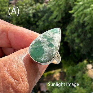 May include: A silver ring featuring a large, teardrop-shaped green gemstone with white marbling. The ring is held up against a blurred green background. The text "Sunlight image" is visible at the bottom.