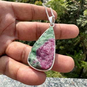 Könnte beinhalten: Eine Anhängerkette mit einem großen, unregelmäßig geformten Stein, der in einer silberfarbenen Lünette gefasst ist. Der Stein zeigt eine Mischung aus grünen und tiefrosa Farbtönen. Die Halskette hat eine zarte Silberkette.
