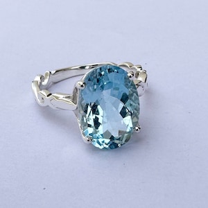 Op de afbeelding: Een zilveren ring met een grote ovale blauwe aquamarijn edelsteen. De ring heeft een delicaat, ingewikkeld ontwerp.