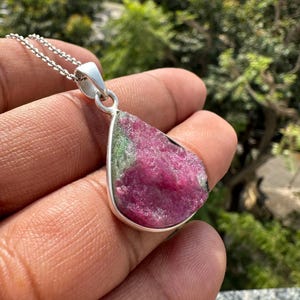 May include: A silver pendant necklace with a teardrop-shaped gemstone, displaying a vibrant mix of pink and green. The pendant is set in a silver bezel and hangs from a delicate silver chain. The natural stone has a rough, textured surface.