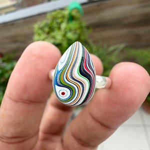 Puede incluir: Un anillo en forma de lágrima con un patrón de capas multicolor que presenta líneas onduladas en blanco, azul, naranja, negro y rojo. La piedra pulida está engastada en una banda de plata lisa, mostrando un aspecto veteado con bandas de colores fluidas.