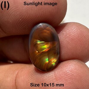 May include: An oval-shaped gemstone with a mix of brown, gold, and green colors. The text "Sunlight image" and "Size 10x15 mm" are visible. The gemstone is held between fingers.