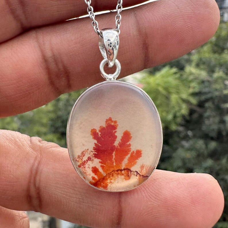 May include: A silver pendant necklace featuring an oval-shaped, translucent stone with a vibrant red and orange dendritic pattern. The pendant hangs from a delicate silver chain, creating a striking contrast.