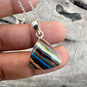 Puede incluir: Un collar de plata con un colgante triangular que presenta una piedra colorida y rayada. La piedra tiene una variedad de colores, incluyendo negro, blanco, rojo, naranja, amarillo, verde y azul.