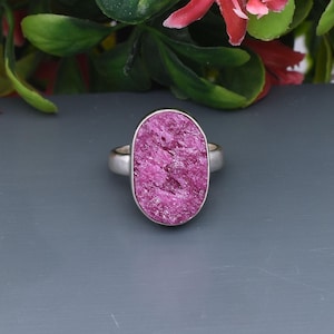 Puede incluir: Un anillo de plata con una gema rosa grande, de forma ovalada y tallada en bruto. La gema tiene una superficie texturizada y está engastada en una sencilla banda de plata. El anillo se muestra sobre un fondo gris con follaje verde y rojo.
