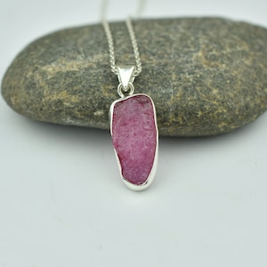 Puede incluir: Un collar con un colgante de plata con una gran piedra preciosa rosa en bruto. La piedra preciosa está engastada en un engaste de plata y cuelga de una delicada cadena de plata.