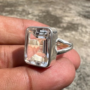 Puede incluir: Un anillo de plata con una gran piedra preciosa rectangular transparente. El anillo tiene un diseño simple y elegante.