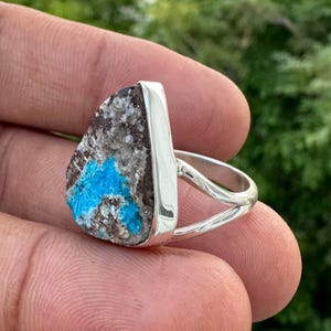 May include: A silver ring featuring a natural stone with a mix of gray, brown, and vibrant turquoise blue hues. The stone has a rough, textured surface and is set in a polished silver band.