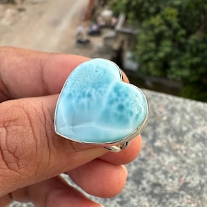 May include: A heart-shaped ring featuring a light blue gemstone with white marbling, set in a silver-colored metal band. The gemstone has a textured appearance, and the ring is held in a hand.
