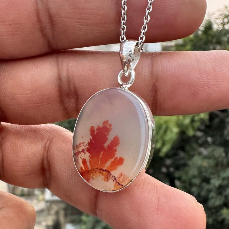 May include: Oval pendant with a silver chain. The pendant features a polished, light-colored stone with a vibrant red and orange, tree-like pattern. The stone is set in a silver bezel.