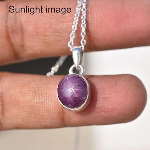 May include: A silver necklace featuring a round, cabochon-cut purple star sapphire pendant with a visible six-rayed star effect. The pendant is set in a simple silver bezel and hangs from a delicate silver chain. The text Sunlight image is visible.
