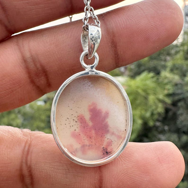 May include: Oval pendant necklace with a silver chain. The pendant features a natural stone with a cream and pink pattern. The stone is set in a silver bezel. The necklace is held by a person's hand.