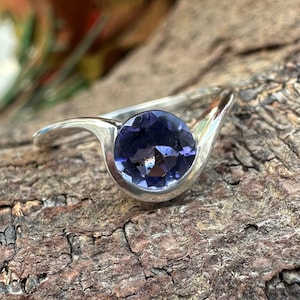 May include: A silver ring featuring a large, round, deep purple gemstone. The ring's band curves gracefully around the stone, creating a modern and elegant design. The gemstone's facets catch the light, showcasing its color and brilliance. The ring is set against a textured wooden surface.