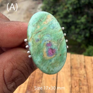 May include: An oval-shaped ring featuring a large, polished green stone with natural brown veining and a prominent magenta-pink inclusion in the center. The stone is set in a silver-toned metal band with small decorative beads, measuring 17x30 mm.
