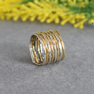 May include: A wide band ring composed of multiple interlocking thin bands in alternating silver and gold tones. Each individual band features a textured, hammered surface with a slightly irregular, wavy edge, creating a layered appearance.
