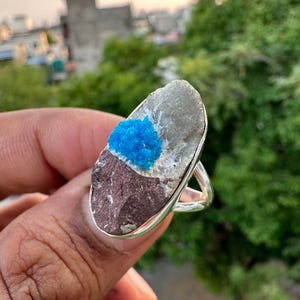 May include: A silver ring featuring a natural stone with a blue crystal cluster. The stone is a mix of gray, brown, and white, set in a simple silver band. The ring is a unique piece of jewelry.