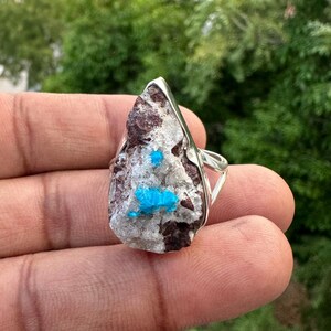 May include: A silver ring featuring a large, irregularly shaped stone. The stone has a mix of colors, including brown, gray, and white, with vibrant blue crystal clusters. The ring's band is a simple, open design.
