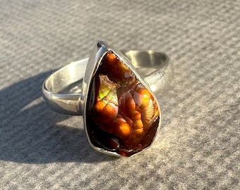 May include: A sterling silver ring featuring a teardrop-shaped gemstone with a mix of brown, orange, and green hues. The gemstone is set in a bezel setting, and the ring has a simple, polished band.