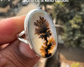 May include: A silver ring featuring an oval-shaped dendritic agate gemstone with a tree-like black and orange pattern. The text (L) see listing for several one of a kind option is visible at the top of the image against a blurred background.