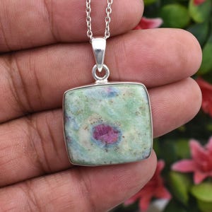 May include: A square pendant necklace featuring a polished, multi-coloured gemstone with a prominent deep pink centre surrounded by mottled light green, white, and blue-grey mineral inclusions, set in a simple silver-toned metal frame on a chain.