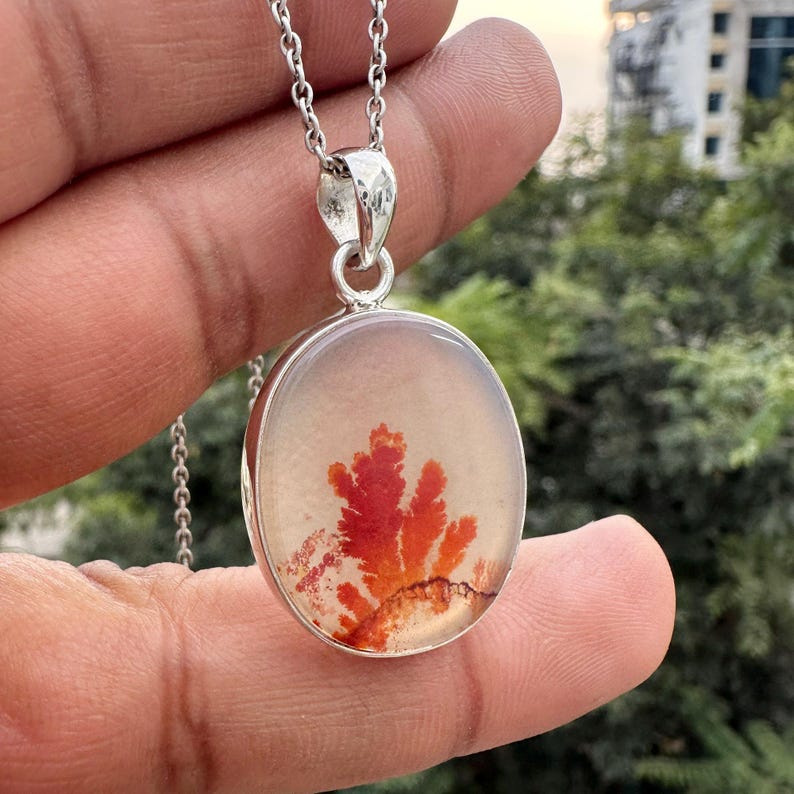 May include: Silver pendant necklace featuring an oval-shaped stone with a red and orange dendritic pattern. The pendant hangs from a delicate silver chain. The stone's design resembles a tree or plant.