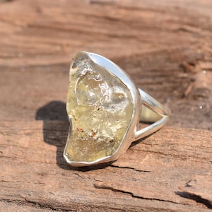 Puede incluir: Un anillo de plata con una gran piedra preciosa de color verde amarillento claro. La piedra preciosa tiene una forma irregular y una textura natural rugosa.