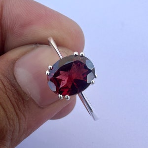 Puede incluir: Un anillo de plata con una gran piedra preciosa roja de forma ovalada. El anillo tiene una banda simple y la piedra preciosa está engastada en garras.