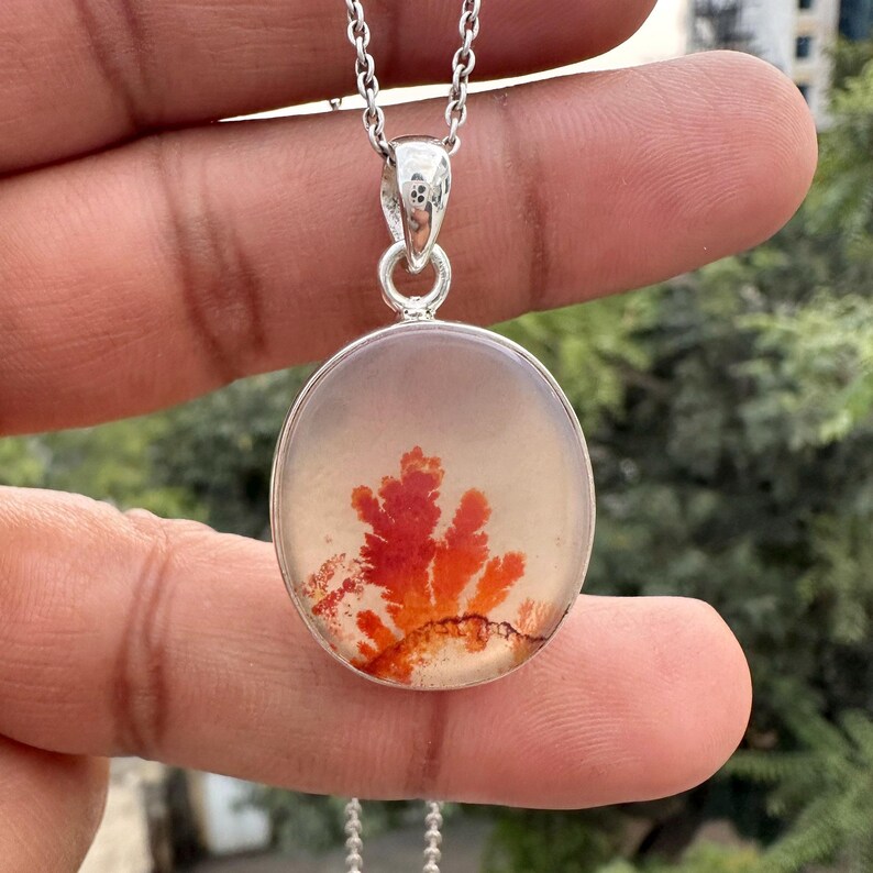May include: A silver pendant necklace featuring a round, clear stone with a vibrant red and orange dendritic inclusion. The pendant hangs from a delicate silver chain, showcasing the natural beauty of the gemstone.