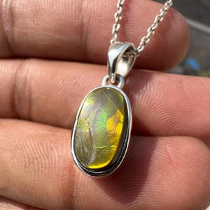 Può includere: Una collana con pendente in argento con una gemma ovale dai colori iridescenti giallo, verde e marrone. La gemma è incastonata in una lunetta d'argento e appesa a una sottile catena d'argento. Il pendente è tenuto in una mano.