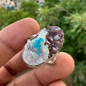 May include: A silver ring featuring a large, oval-shaped stone. The stone has a textured, brown and white surface with a cluster of bright blue crystals. The ring is a unique piece of jewelry.