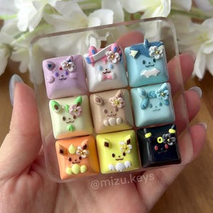 May include: A set of nine square mechanical keyboard keycaps arranged in a clear plastic grid, each featuring a unique sculpted character face with floral accents in pastel colours, including purple, white, blue, green, brown, orange, yellow, and black.