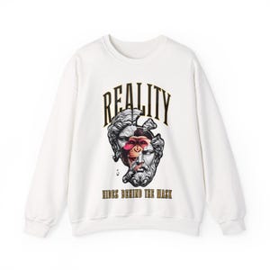 Puede incluir: Sudadera blanca de cuello redondo con un diseño gráfico. El diseño presenta una cara dividida de una estatua clásica y un mono, con la palabra "REALITY" arriba y "HIDES BEHIND THE MASK" abajo.