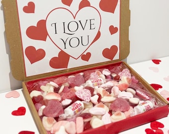 May include: A cardboard box with a red interior filled with assorted pink and white candies. The box lid features a white heart with the words "I LOVE YOU" and red heart accents. Valentine's Day themed.