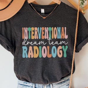 May include: Dark grey heather t-shirt with the text "Interventional Dream Team Radiology" in colorful letters. The shirt is being modeled on a person wearing blue jeans and a brown hat.