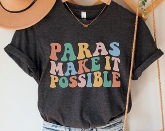 May include: Dark grey heather V-neck t-shirt with the text "Paras Make It Possible" in rainbow colors. The shirt is on a hanger with a brown hat and a woven bag.