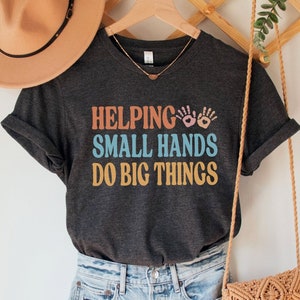 May include: Dark grey heather t-shirt with the text "Helping Small Hands Do Big Things" in colorful letters with handprint graphics.