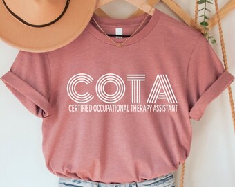 May include: A heather mauve t-shirt with the text "COTA Certified Occupational Therapy Assistant" in white. The shirt is on a hanger with a straw hat and a woven bag.