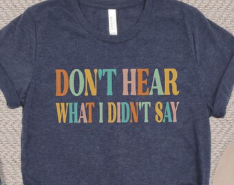 May include: Heather navy blue t-shirt with the text "DON'T HEAR WHAT I DIDN'T SAY" in orange, pink, and teal lettering. The shirt is styled with rolled-up sleeves and a knot at the bottom. A pair of blue jeans and a hat are partially visible.