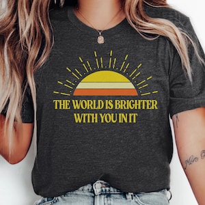 May include: Dark grey heather t-shirt featuring a graphic of a rising sun with yellow, orange, and white stripes. The text "THE WORLD IS BRIGHTER WITH YOU IN IT" is printed below the sun graphic.