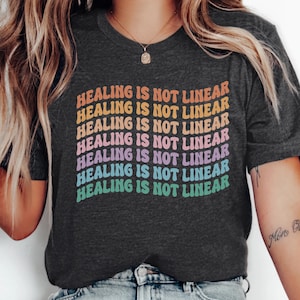 May include: Dark grey heather t-shirt with the text "HEALING IS NOT LINEAR" in a rainbow gradient. The text is stacked vertically. The shirt is a crew neck style.