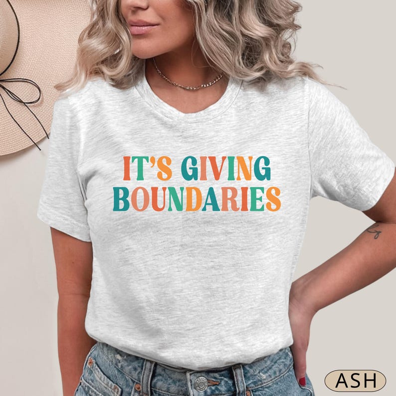 May include: A light gray t-shirt with the text "IT'S GIVING BOUNDARIES" in teal, orange, and green. The shirt is short-sleeved and has a crew neck. The word "ASH" is printed in a small circle in the bottom right corner.