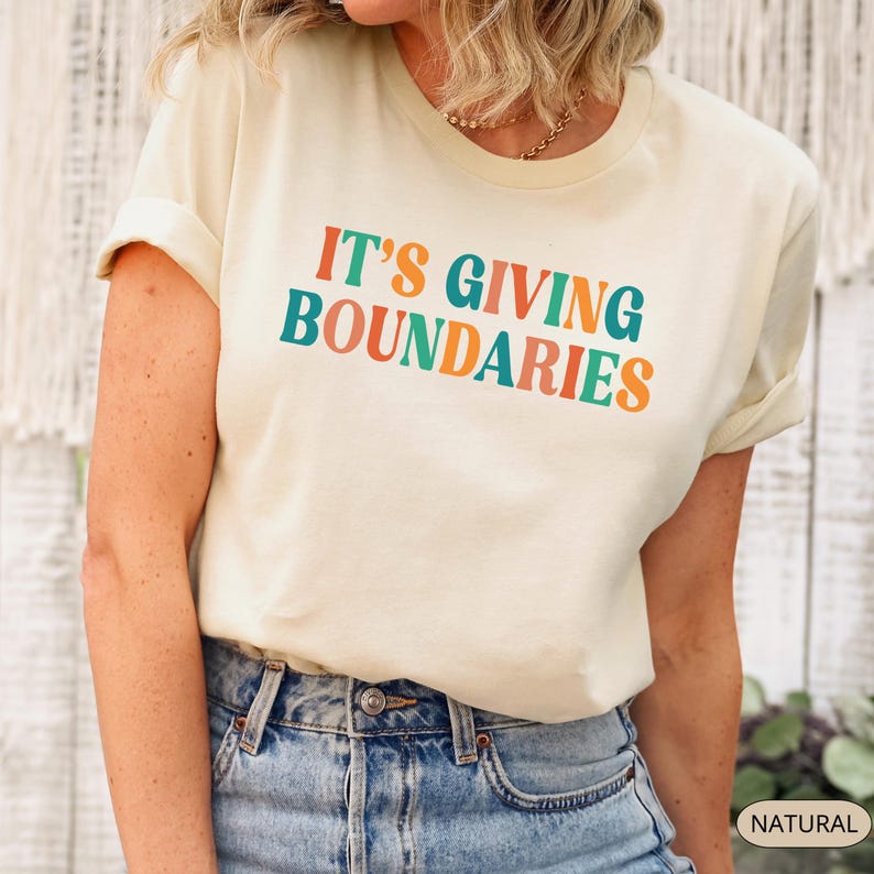 May include: Cream-colored t-shirt with the text "IT'S GIVING BOUNDARIES" in orange, teal, and red lettering. The shirt has rolled-up sleeves and is paired with blue jeans. The word "NATURAL" is visible in the bottom right corner.