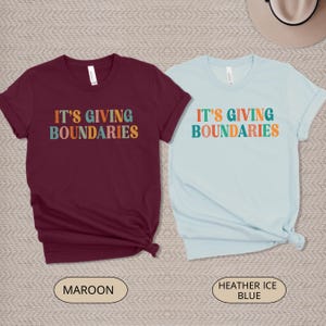 May include: Two short-sleeved t-shirts, one maroon and one light blue. Each shirt features the text "IT'S GIVING BOUNDARIES" in colorful, retro-style lettering. The maroon shirt has a "Maroon" label, and the blue shirt has a "Heather Ice Blue" label.