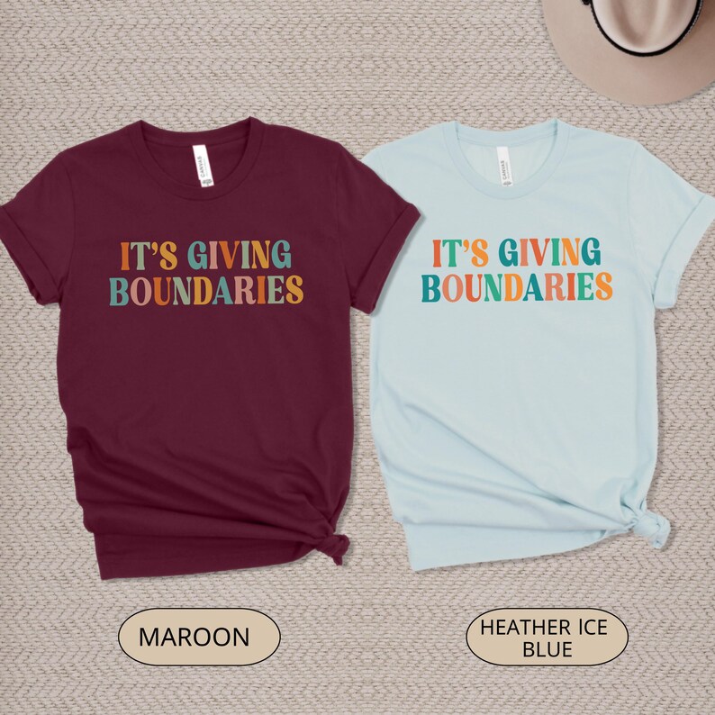May include: Two short-sleeved t-shirts, one maroon and one light blue. Each shirt features the text "IT'S GIVING BOUNDARIES" in colorful, retro-style lettering. The maroon shirt has a "Maroon" label, and the blue shirt has a "Heather Ice Blue" label.